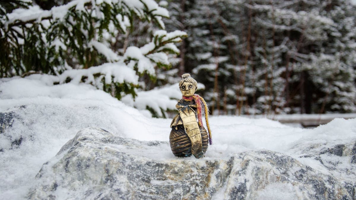 A quirky figurine stands on a rock amidst a serene, snow-covered forest landscape.